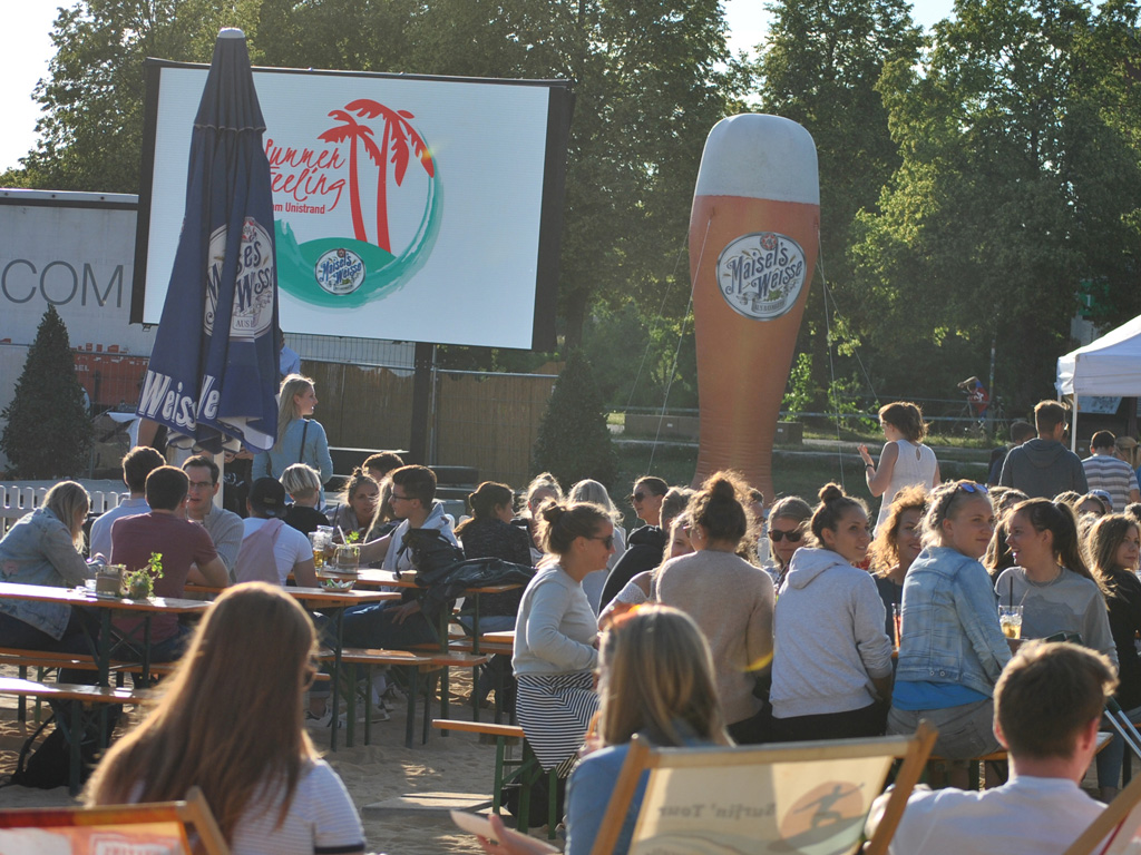 Menschen sitzen gemütlich an Biertischen am Unistrand zusammen