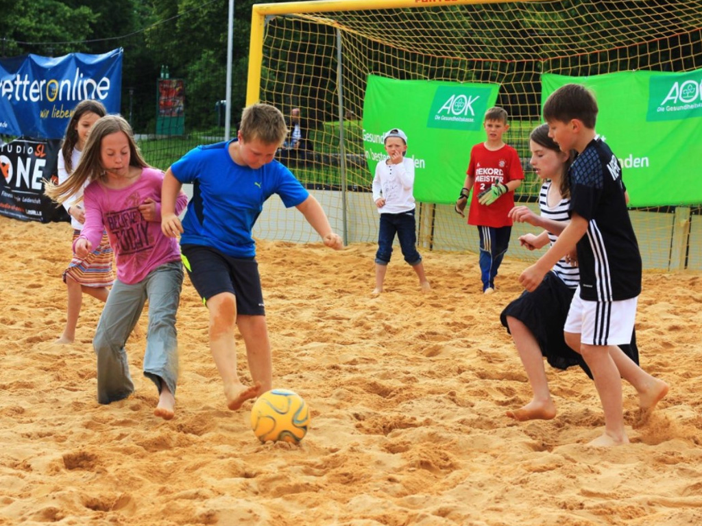 Acht Kinder spielen auf einem Sandplatz Beachsoccer